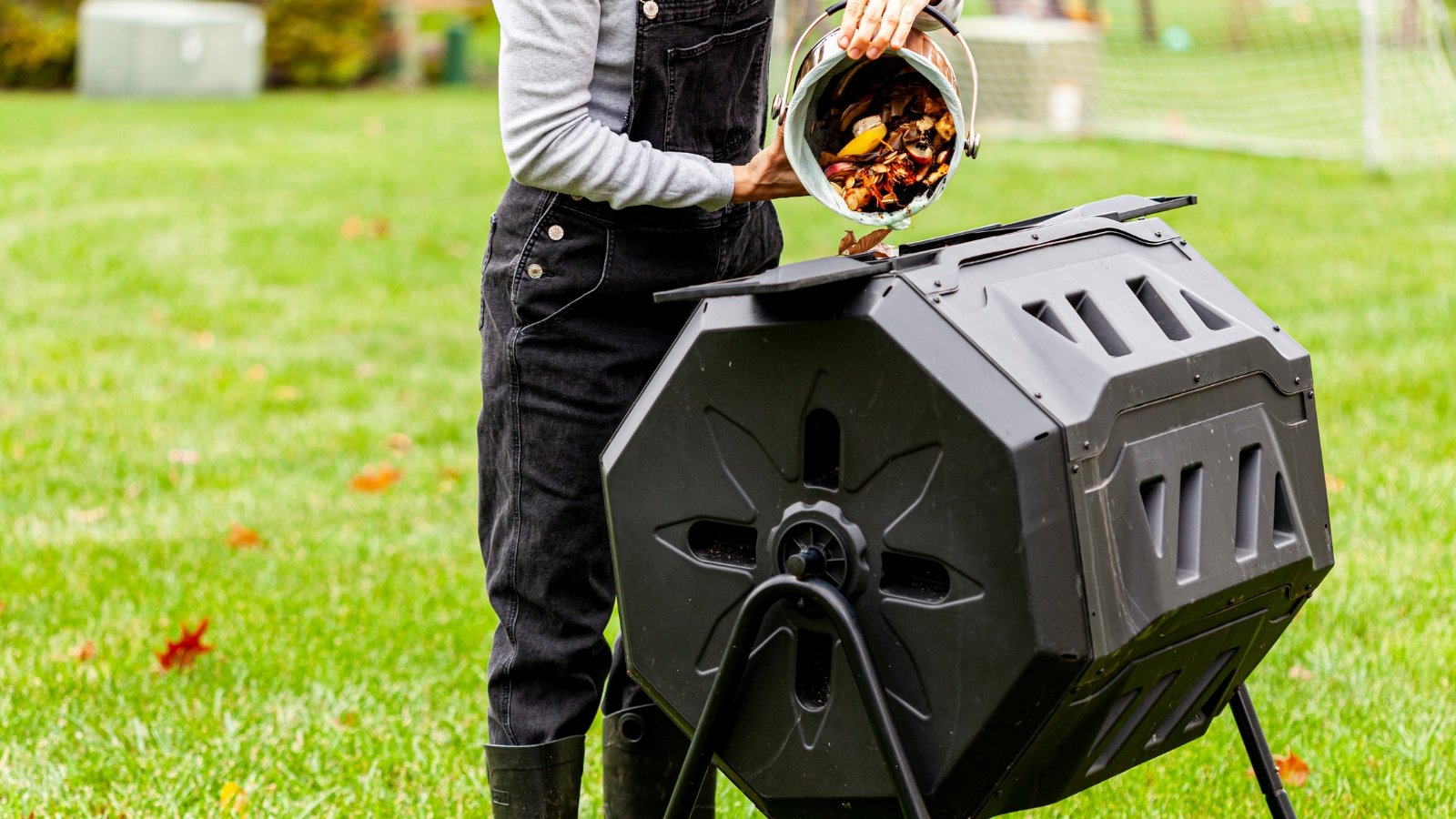 The image depicts a woman tipping a small plastic bin to release a mixture of leftover vegetables and other biodegradable waste into a larger tumbling container intended for soil enrichment purposes.