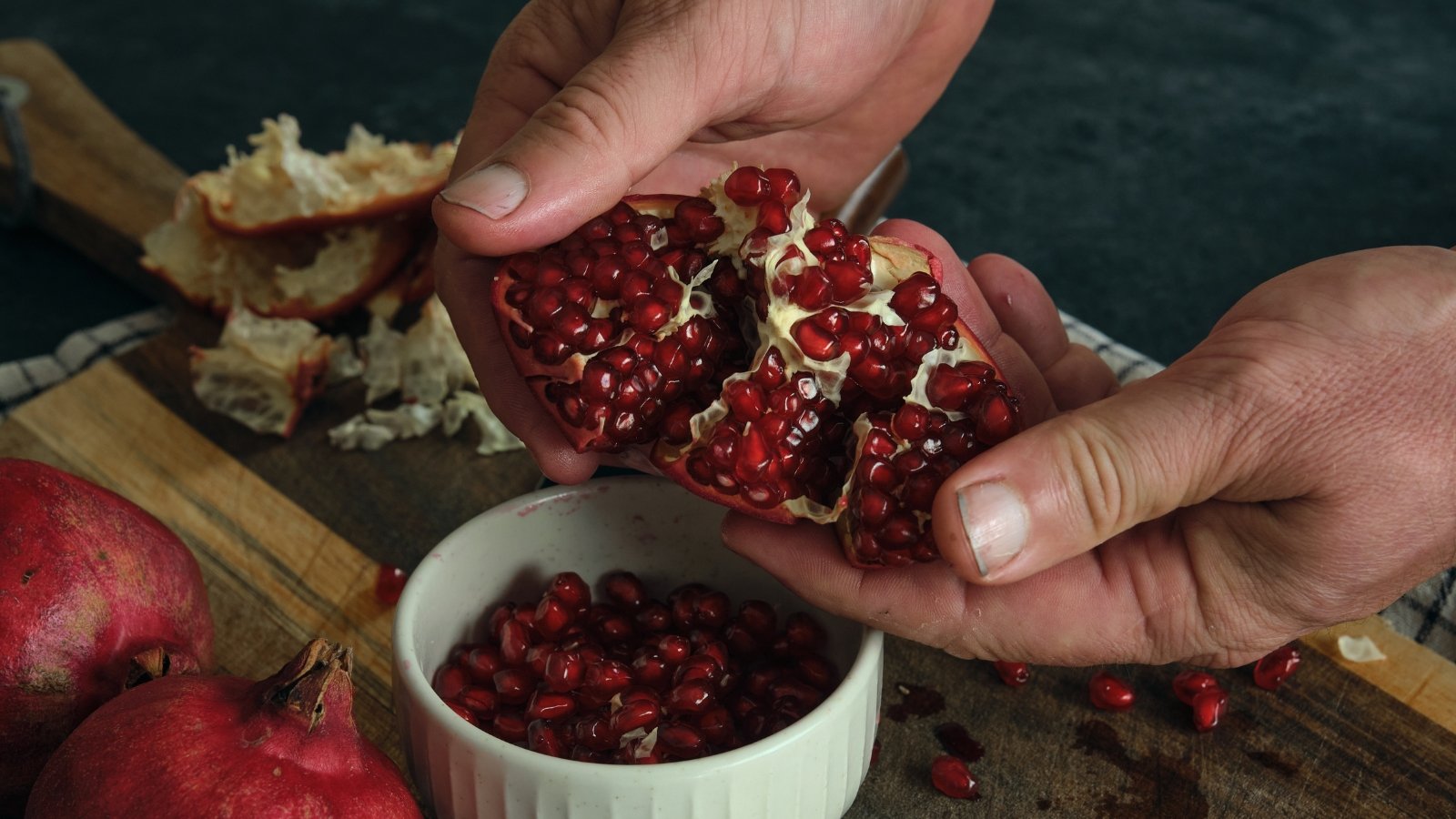 The man breaks open a ripe red pomegranate, revealing its glistening, jewel-like seeds and juicy, ruby-red pulp spilling out.